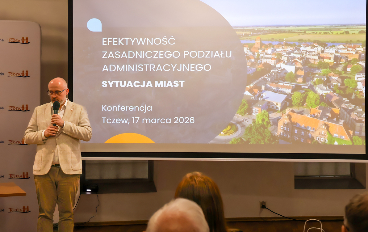 Zdjęcie przedstawia mężczyznę z mikrofonem stojącego przy banerze z nazwą wydarzenia