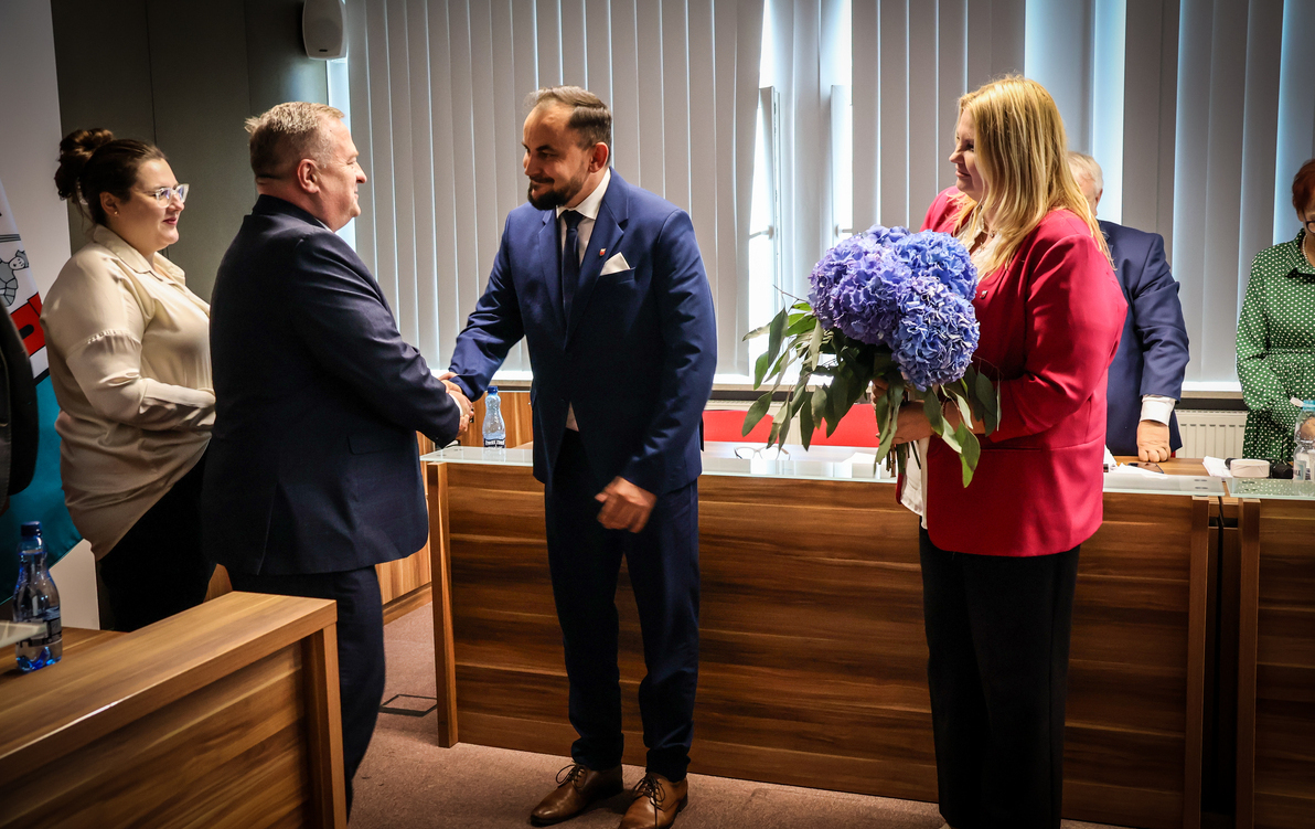 Zdjęcie przedstawia dwóch mężczyzn podających sobie dłonie. Z boku stoi kobieta z bukietem kwiatów