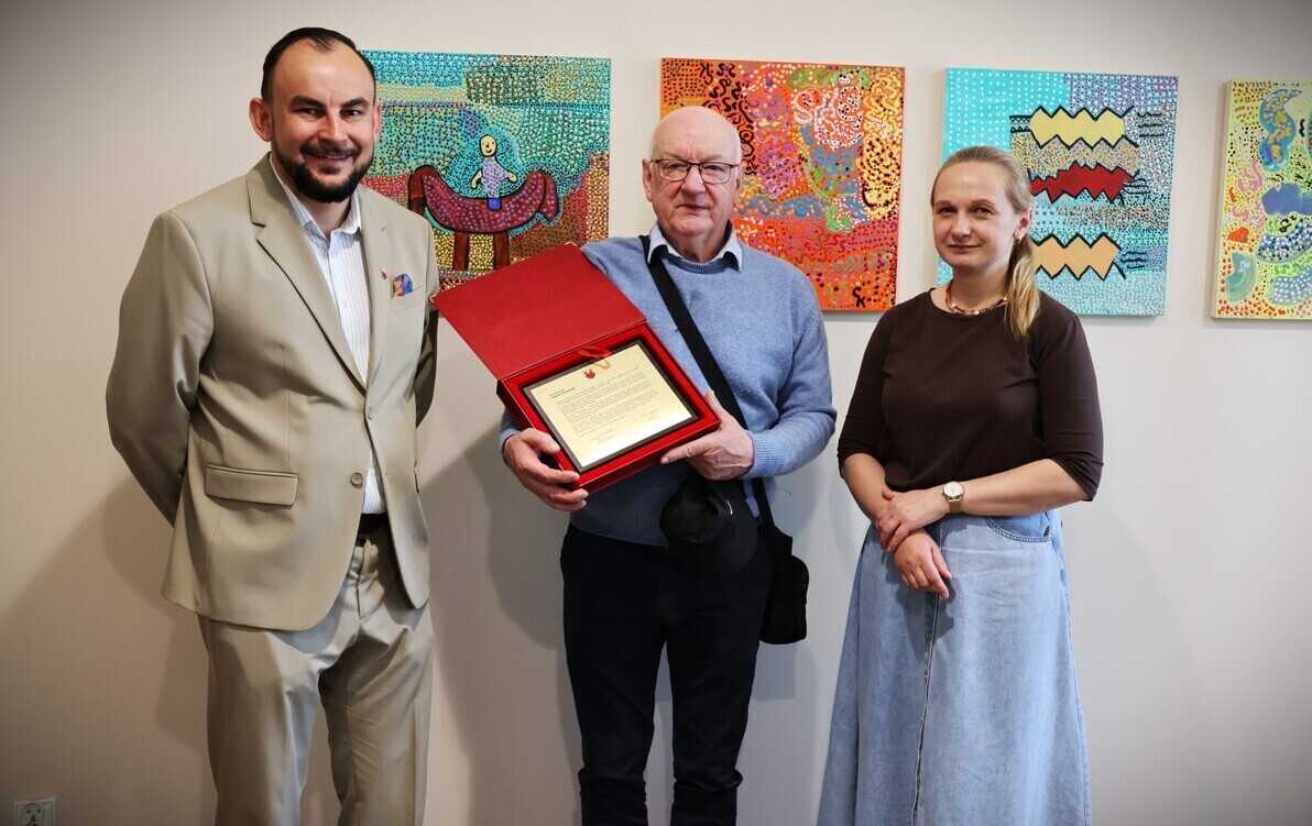 Zdjęcie przedstawia trzy osoby stojące na tle ściany z kolorowymi obrazami. Mężczyzna w środku trzyma w ręku grawerton gratulacyjny