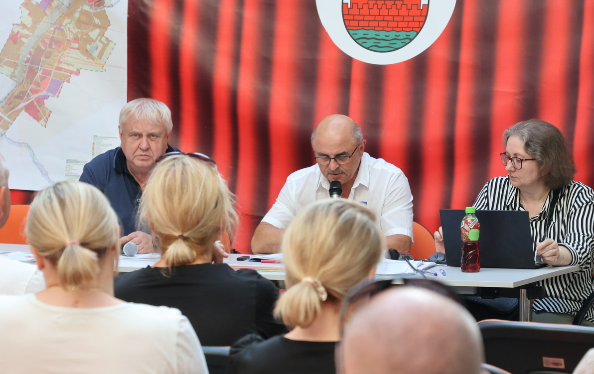 Zdjęcie przedstawia trzy osoby siedzące za stołem i kilka słuchających ich osób, siedzących tyłem