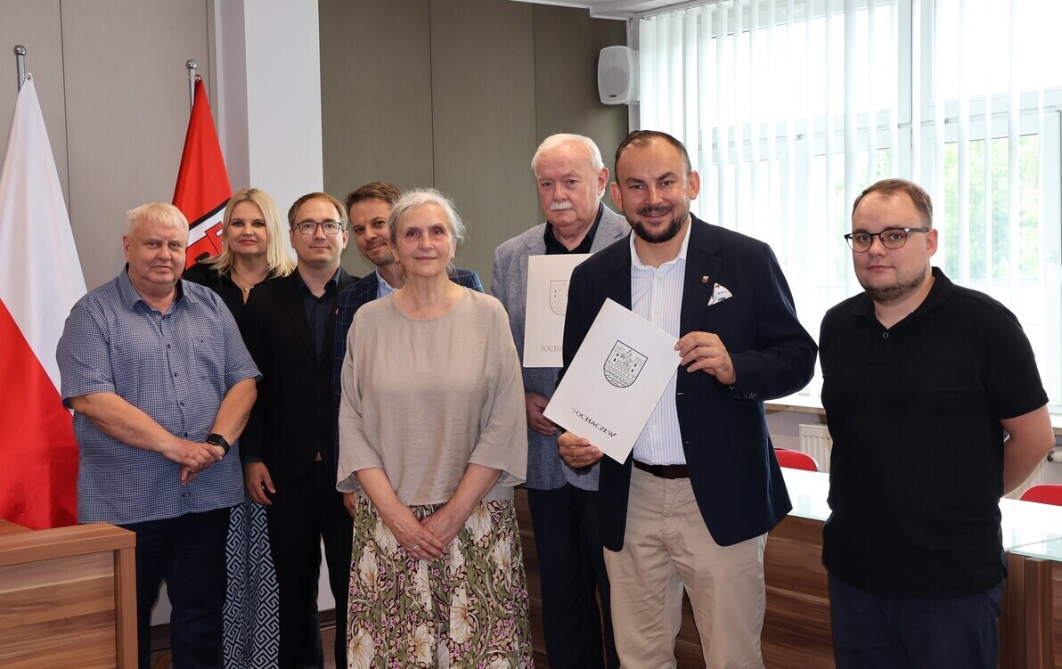 Zdjęcie przedstawai grupę osób stojących na tle ściany i polskiej flagi