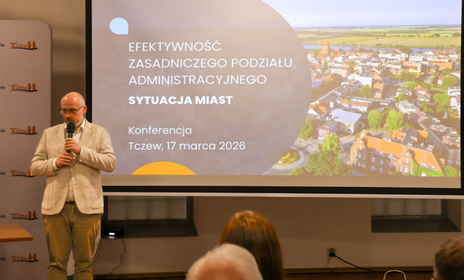 Zdjęcie przedstawia mężczyznę z mikrofonem stojącego przy banerze z nazwą wydarzenia