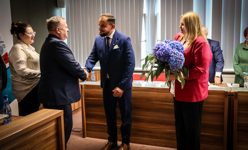 Zdjęcie przedstawia dwóch mężczyzn podających sobie dłonie. Z boku stoi kobieta z bukietem kwiatów