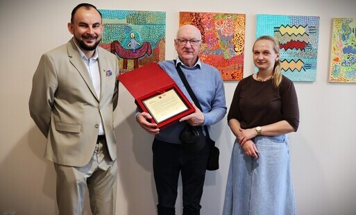 Zdjęcie przedstawia trzy osoby stojące na tle ściany z kolorowymi obrazami. Mężczyzna w środku trzyma w ręku grawerton gratulacyjny