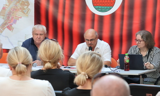 Zdjęcie przedstawia trzy osoby siedzące za stołem i kilka słuchających ich osób, siedzących tyłem