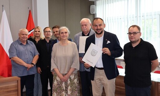 Zdjęcie przedstawai grupę osób stojących na tle ściany i polskiej flagi